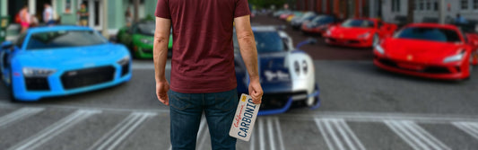Man holding his front license plate in hand and standing in front of cars at a car show